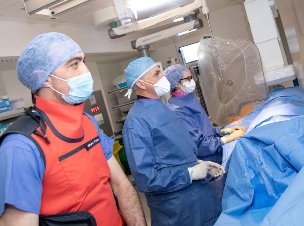 A team of three cardiologists in scrubs perform a Cath Scan in a theatre room. Patient is gowned and masked lying on table.